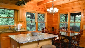 A wooden cabin interior with a kitchen featuring granite countertops, a dining table with chairs, and large windows offering a view of the forest.