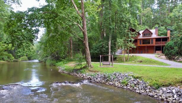 A riverside lodge with a wooden deck and a swing chair, surrounded by lush greenery.