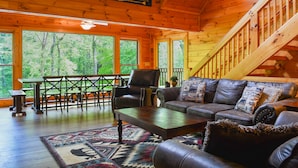 A cozy cabin interior with a wooden staircase, a leather sofa, a dining table with chairs, and large windows offering a view of the forest.
