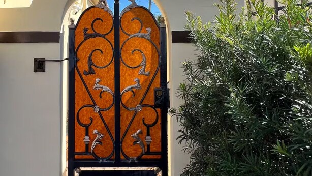 A wrought iron gate with intricate designs, leading to a building with a white facade and an arched entrance. - Luxurious little home! (Hialeah)