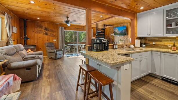 Un intérieur de chalet chaleureux avec une cheminée, des sièges confortables, une cuisine dotée de plans de travail en granit et une grande fenêtre donnant sur un paysage verdoyant.