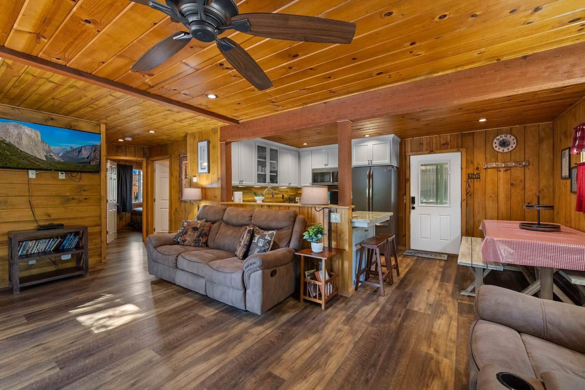 A spacious living room with a large sofa, a ceiling fan, a kitchen area with white cabinets, and a dining table with a checkered tablecloth.