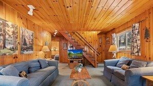 A cozy cabin interior with two sofas, a wooden coffee table, a TV, and a staircase leading to a loft.