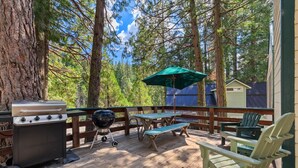 A wooden deck with a barbecue grill, picnic table, and chairs, surrounded by tall trees.