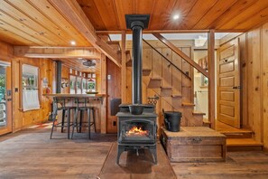 A cozy cabin interior with a wood-burning stove, wooden walls, a bar area, and a staircase.