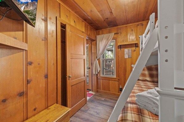 A wooden cabin interior with a bunk bed, a wooden door, and a window with curtains.