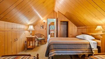 A cozy attic bedroom with wooden walls and a sloped ceiling. It features a bed with a striped bedspread, a bedside table with a lamp, and a chair.