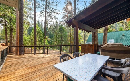 A wooden deck with a table and chairs, surrounded by tall trees and a green fence.