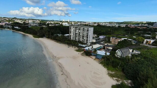 Luchtbeeld van een kustgebied met een zandstrand, een groot hotelgebouw en omliggende woongebouwen.