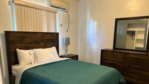 Een keurig opgemaakt bed met een blauwgroene sprei en witte kussens, een bedlamp, een houten dressoir met spiegel, en een wandairconditioner.