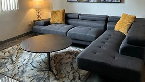 Een moderne woonkamer met een hoekbank, een ronde salontafel en een groot abstract schilderij aan de muur.