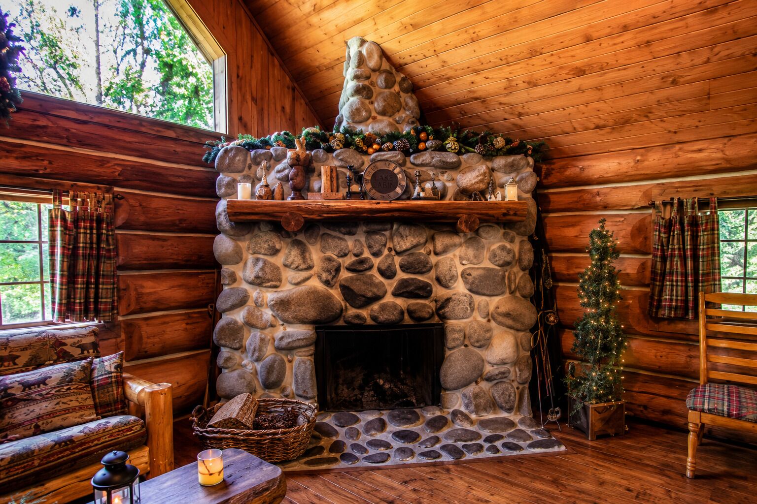 A log cabin interior with a stone fireplace, plaid curtains, a Christmas tree, and a cozy seating area.