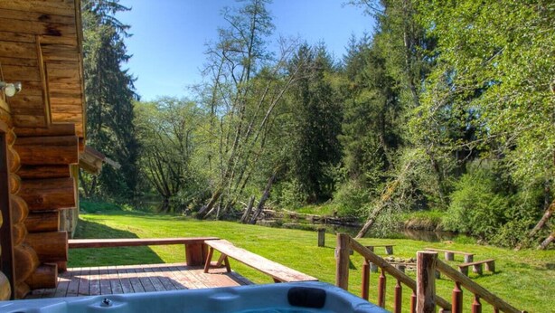 A hot tub with a wooden deck and picnic tables, surrounded by a wooden fence and lush greenery.