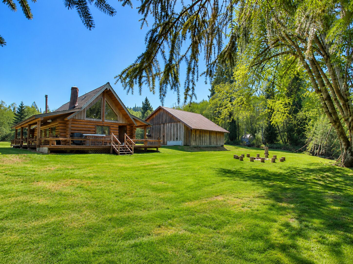 A log cabin with a porch and a separate building, surrounded by a well-maintained lawn and trees.