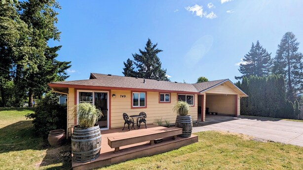 A single-story house with a number 749 on the front, a covered patio with a table and chairs, and two large planters.