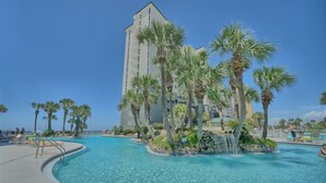 A pool area with a waterfall feature, surrounded by palm trees and a tall building in the background.