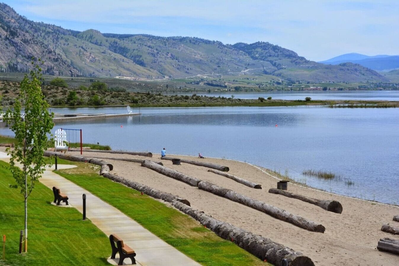 A lakeside park with a walking path, benches, and a view of distant hills.