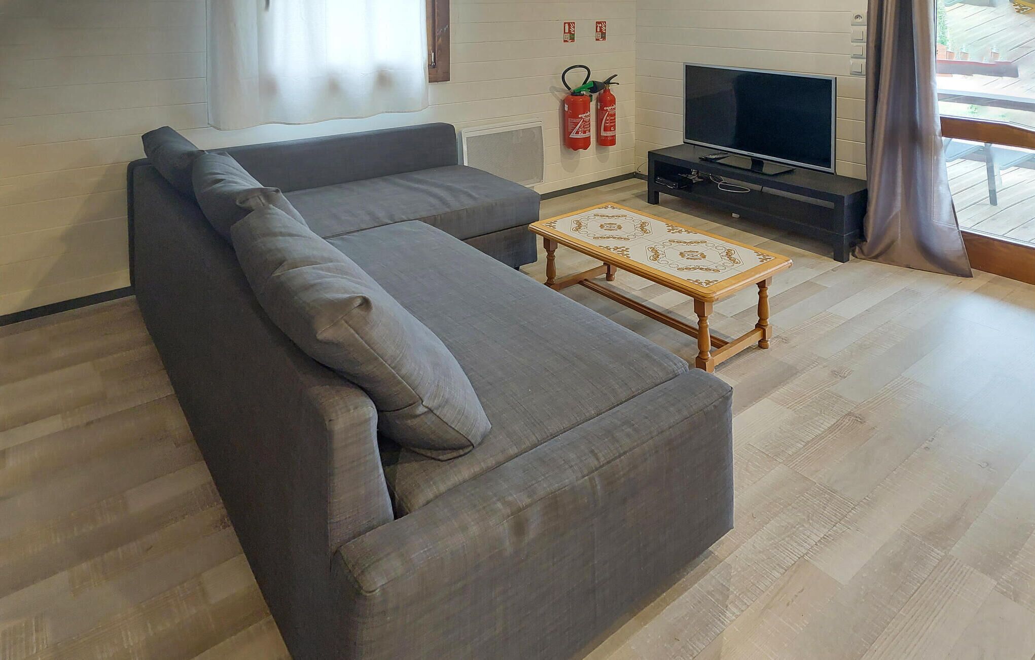 Un salon moderne avec un canapé d’angle gris, une table basse en bois, un téléviseur à écran plat et des extincteurs fixés au mur.
