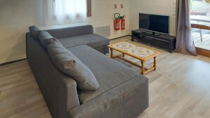 Un salon moderne avec un canapé d’angle gris, une table basse en bois, un téléviseur à écran plat et des extincteurs fixés au mur.