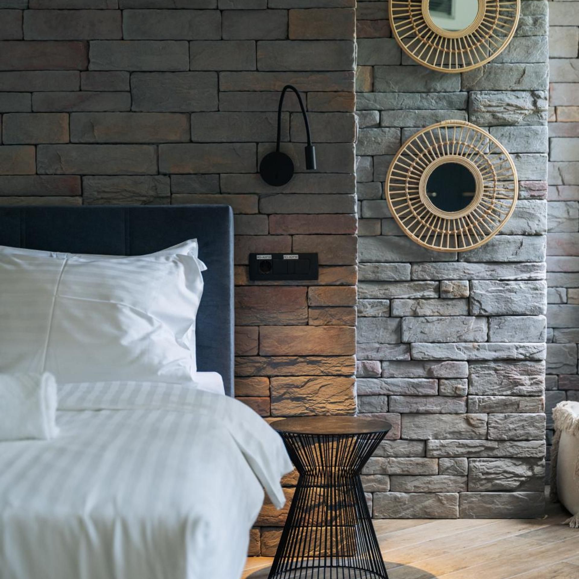 Un dormitorio con pared de piedra, una cama con ropa blanca, una mesita pequeña y dos espejos montados en la pared.