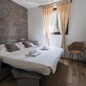 A hotel room with a bed, a chair, a window with curtains, a brick wall, and a radiator. - Rezidenca Ortus (Ankaran)