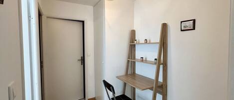 A small, well-lit room with a wooden floor, a black folding chair, a wooden ladder shelf, and a door.