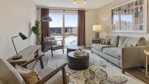 A modern living room with a sofa, armchair, and coffee table, a large window with curtains, and a city view. - 5* Luxury, City Centre, Brand New Development (Belfast)
