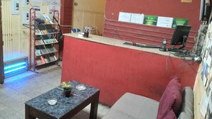 Una zona de recepción con un mostrador rojo, relojes mostrando diferentes husos horarios, una pequeña mesa con una planta y un área de estar con un sofá.