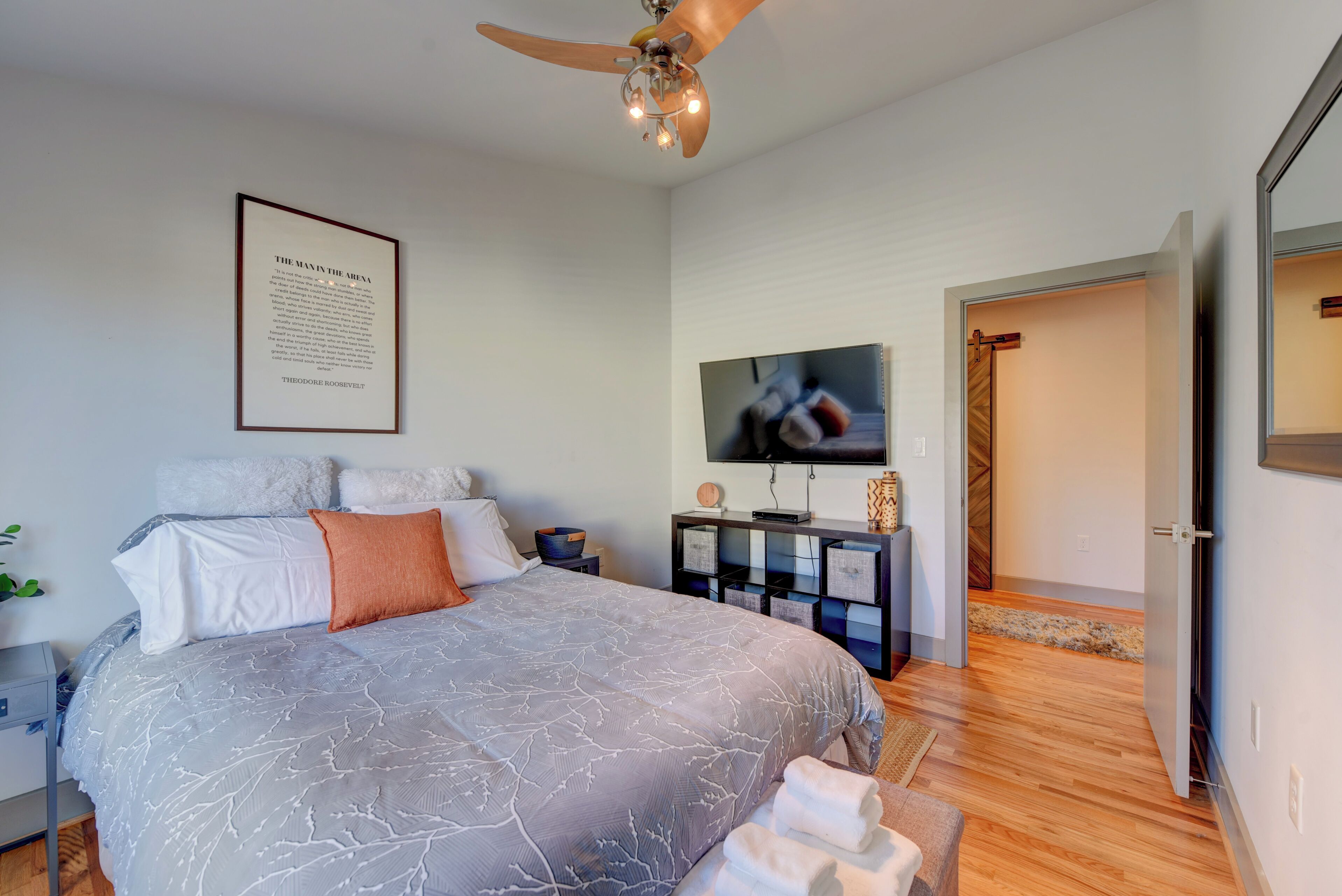 A modern bedroom with a large bed, a ceiling fan, a framed quote on the wall, a TV, and a wooden floor.