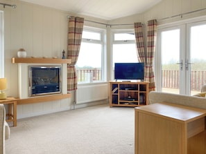 A living room with a fireplace, a television on a wooden stand, and a view of the outdoors through large windows.