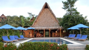 A pool area with a thatched-roof structure, blue lounge chairs, and red flowering bushes. - Lodge Ylang Ylang (Cóbano)