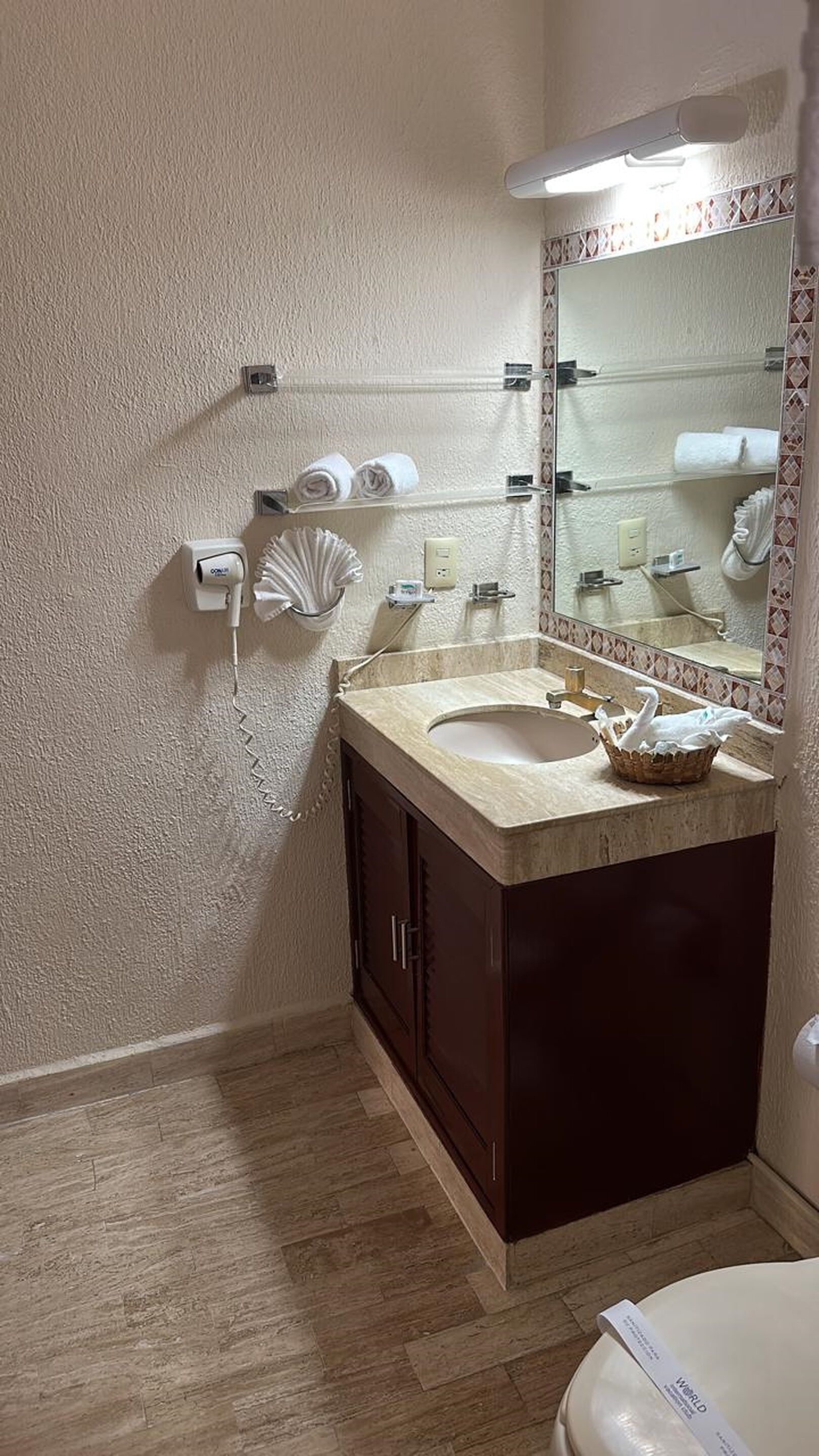 Un baño con un lavamanos color beige, armarios de madera oscura, un espejo con un marco decorativo y un perchero con toallas dobladas.