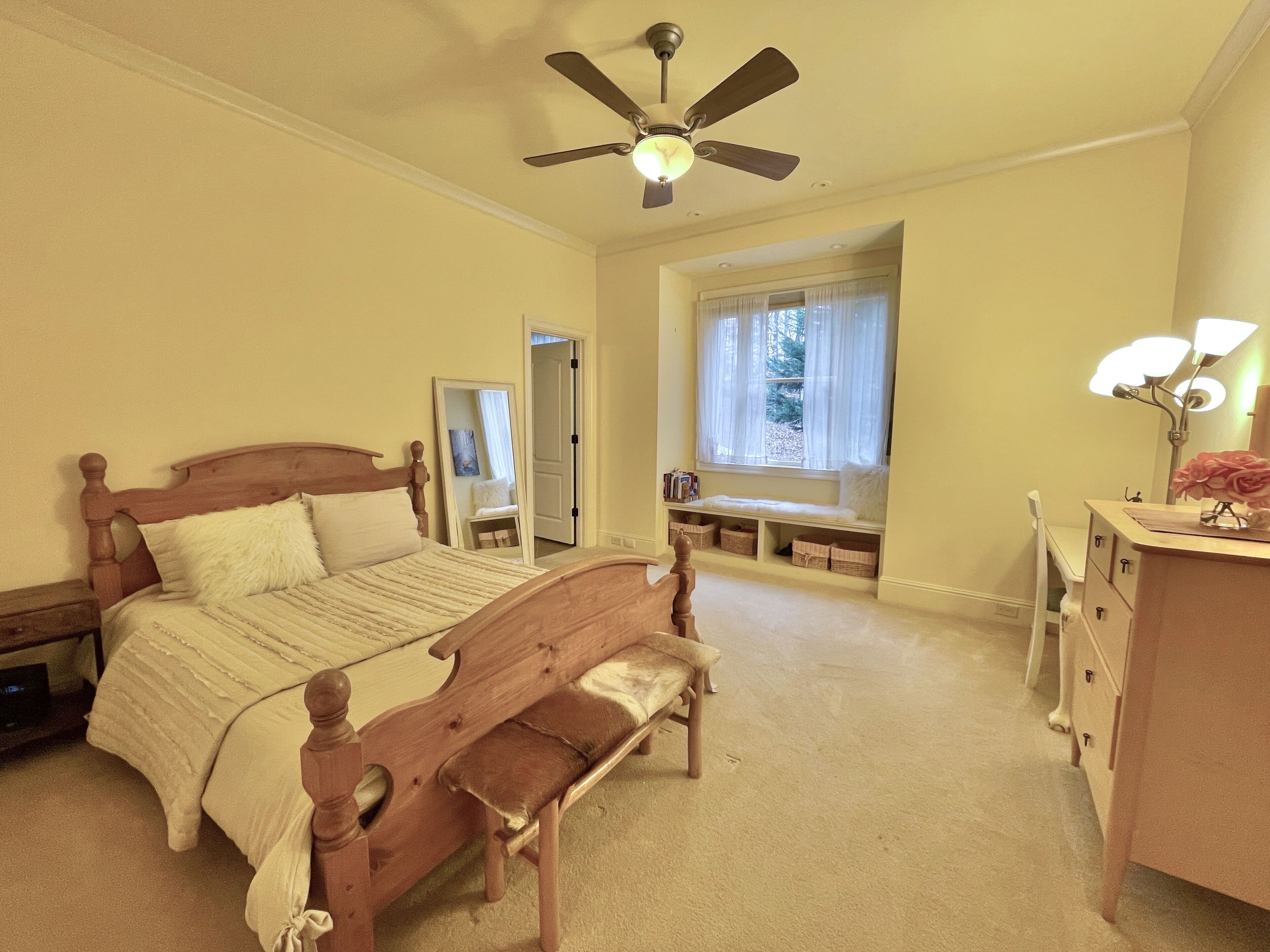 A bedroom with a bed, a bench, a dresser, a mirror, a ceiling fan, and a window with a view of trees.