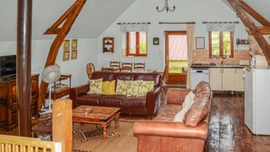 Uma sala de estar acolhedora com um sofá de couro marrom, uma mesa de centro de madeira e um espaço de refeições com cadeiras de madeira. Uma cozinha com geladeira branca e armários de madeira é visível ao fundo.