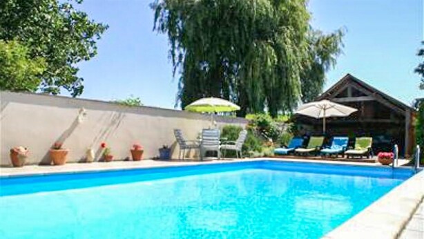 Piscina com espreguiçadeiras e guarda-sóis, cercada por plantas em vasos e um edifício com telhado inclinado.
