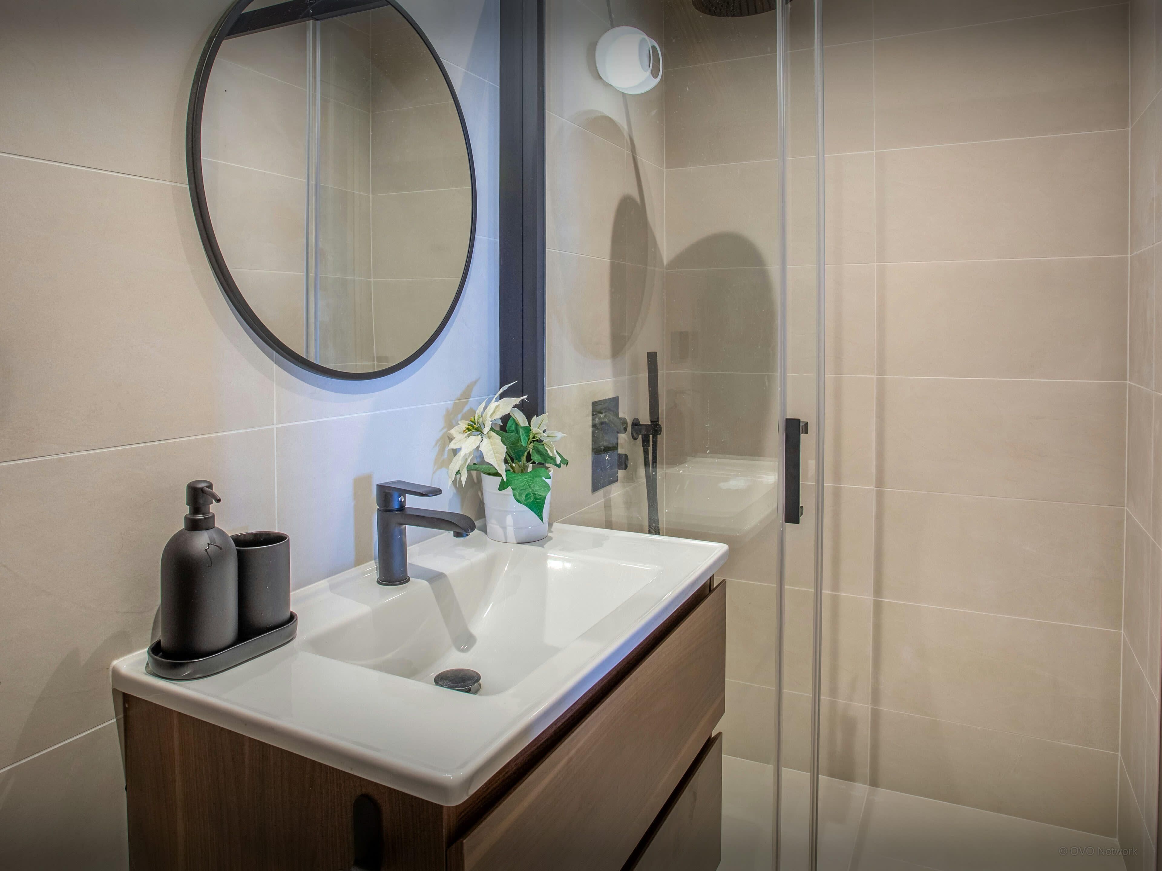 A modern bathroom with a white sink, a round mirror, a potted plant, and a glass shower enclosure.