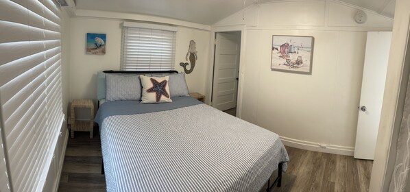 A bedroom with a bed, a small table, a chair, a window with blinds, and a door. - Bayside Cottage - Steps from the Bay and Ocean with AC (San Diego)