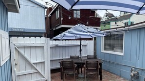 A patio with a table and chairs, a white picket fence, and a blue house. - Bayside Cottage - Steps from the Bay and Ocean with AC (San Diego)
