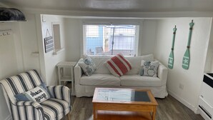 A living room with a striped armchair, a white sofa with patterned cushions, a wooden coffee table, and a window with blinds. - Bayside Cottage - Steps from the Bay and Ocean with AC (San Diego)