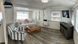 A compact living space with a white sofa, striped armchair, and a wooden coffee table. A dark cabinet is against the wall, and a flat-screen TV is mounted above it. - Bayside Cottage - Steps from the Bay and Ocean with AC (San Diego)