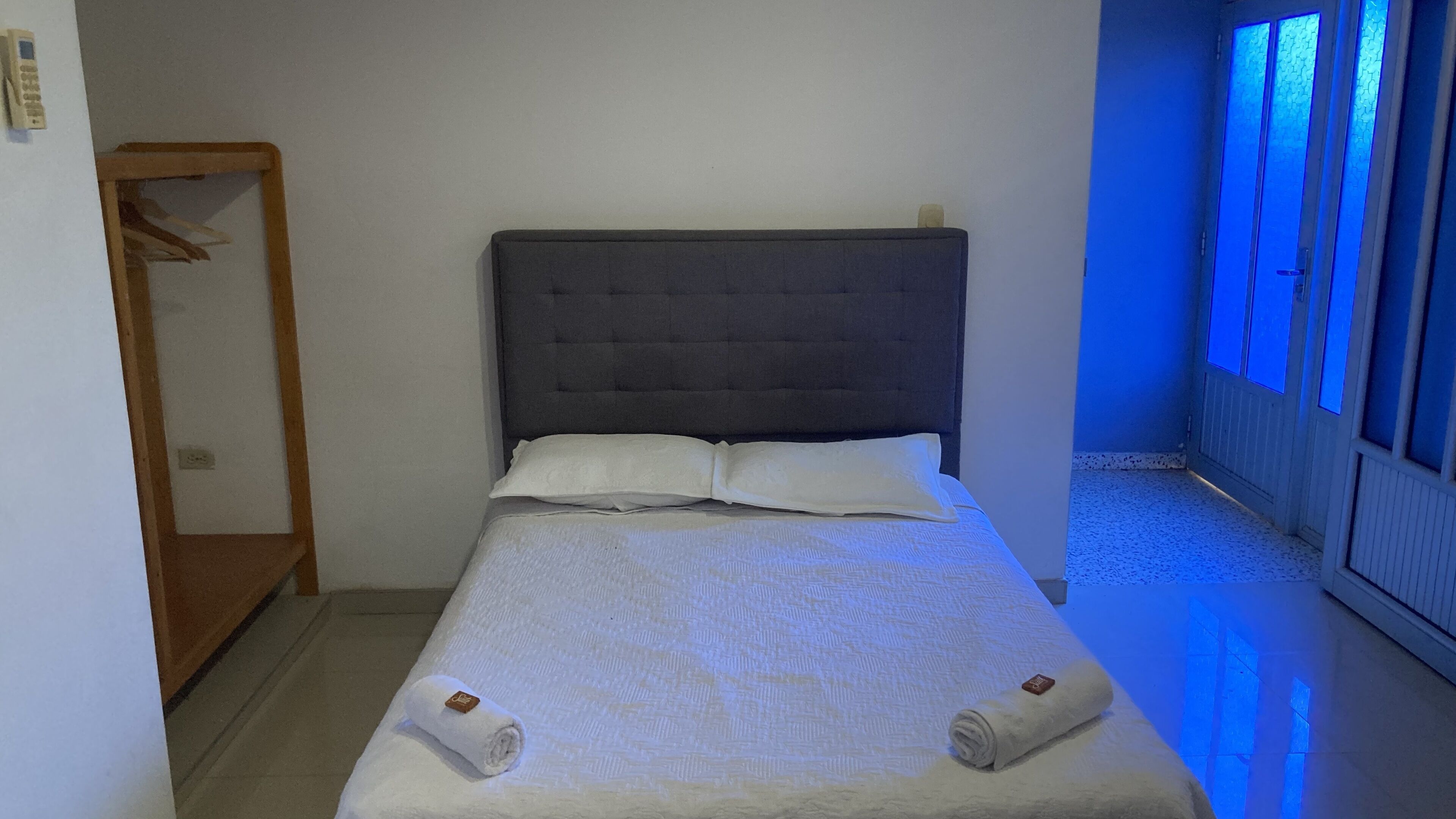 Una cama individual con cabecera gris, ropa de cama blanca y dos toallas enrolladas.