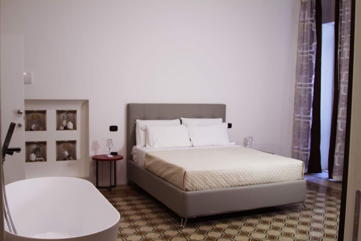 Quarto com cama, mesa de cabeceira, prateleira na parede com objetos decorativos e banheiro com banheira.
