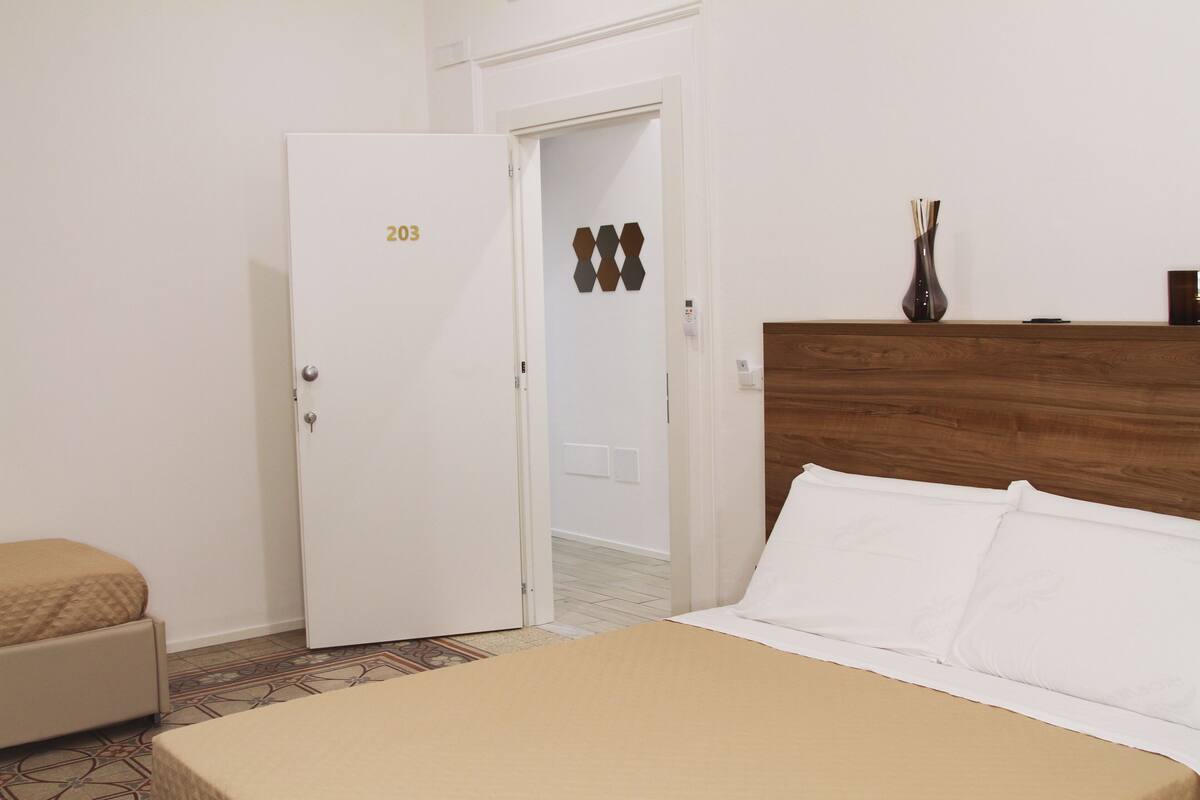 Quarto de hotel com uma cama, uma porta com o número 203, cabeceira de madeira, mesa de cabeceira e um vaso.