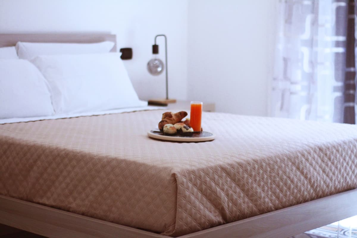 Cama bem arrumada com cobertura acolchoada, bandeja com croissants e um copo de suco de laranja, e uma luminária de cabeceira.
