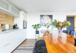 A modern kitchen with white cabinets, a wooden countertop, and a coffee machine. There is a dining table with chairs and a vase of flowers.