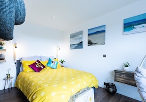 A bedroom with a bed covered in yellow bedding, a wooden headboard, and a small bedside table with a plant. There are two wall-mounted paintings depicting a beach scene.