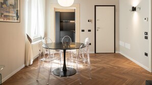 Ein moderner Essbereich mit einem runden Tisch und transparenten Stühlen, einer großen Pendelleuchte und Blick in eine Küche durch eine offene Tür.