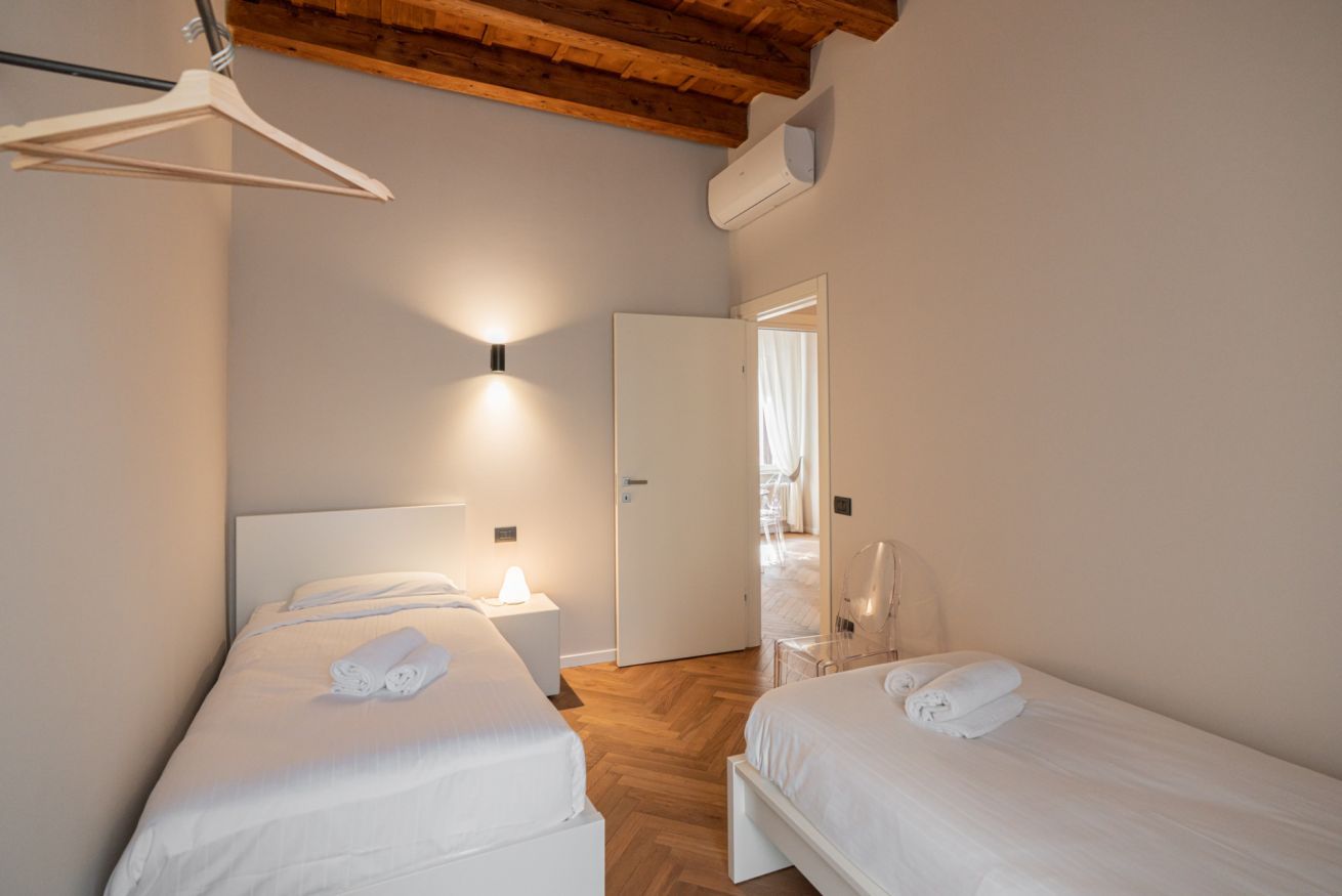 Una camera da letto con due letti, soffitto in legno, parete bianca e porta che dà su un bagno.