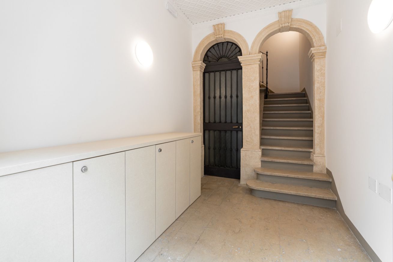 A hallway with a staircase leading to a dark door, white walls, and a light fixture above the door.