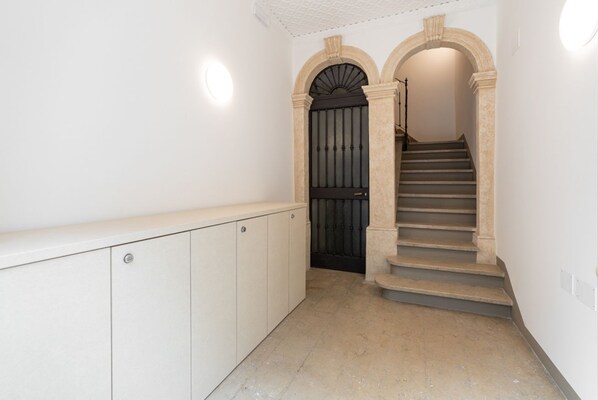 Un pasillo con una escalera que lleva a una puerta oscura, paredes blancas y una lámpara sobre la puerta.